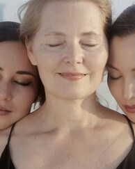 Portrait rapproché de trois femmes, une aînée au centre et deux plus jeunes joue contre joue, yeux clos, dans un geste de tendresse intergénérationnelle